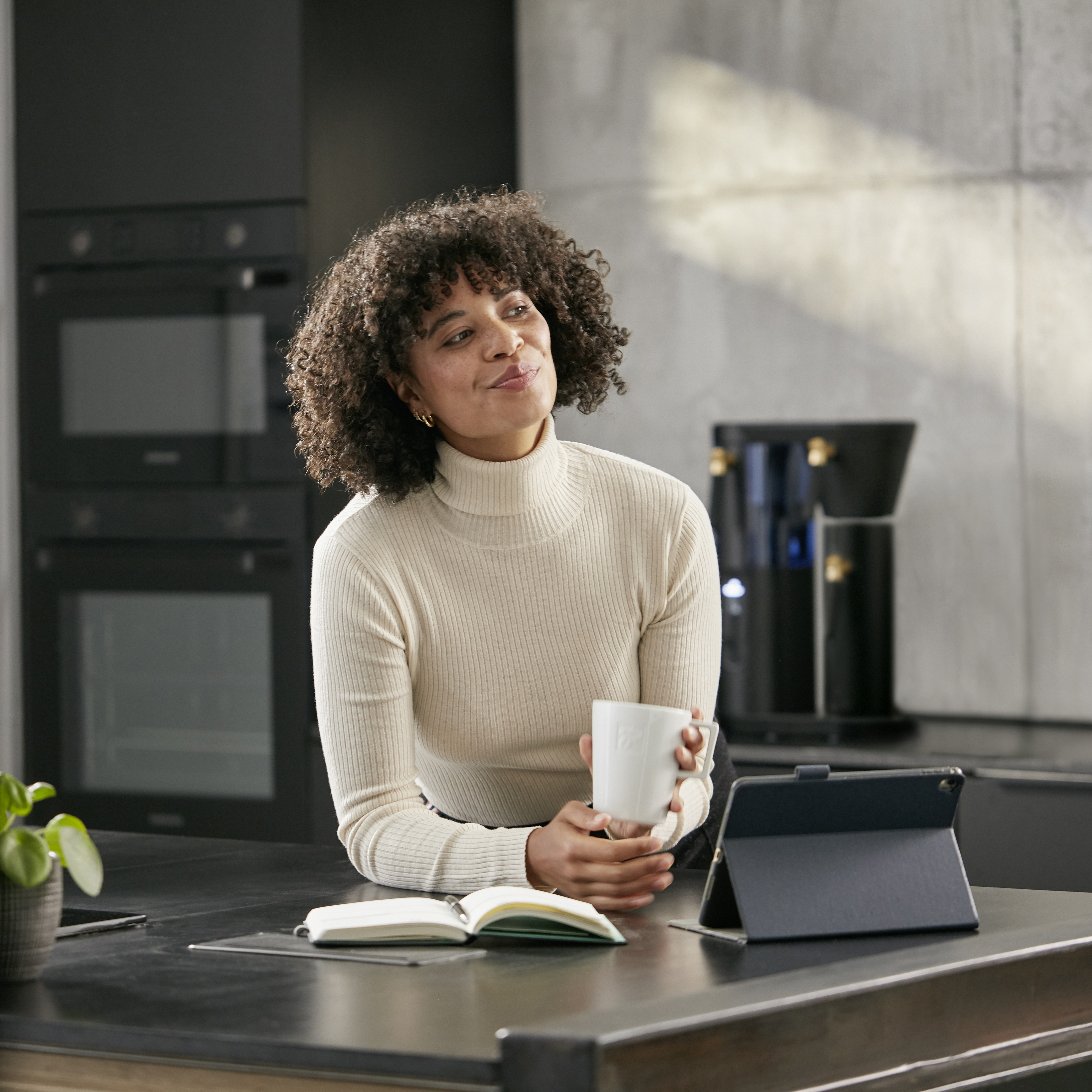 Eine Frau mit einer Tasse in der Hand, die in die Ferne lächelt. Im Hintergrund eine Küche und eine schwarze Filbii.